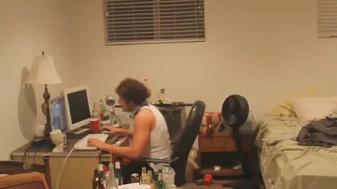 Guy frantically typing at computer in messy dark room late at night, tank top, intense focus, chaos energy
