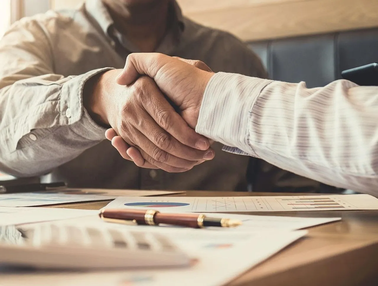 Two business people shaking hands over a table with financial documents and a pen.