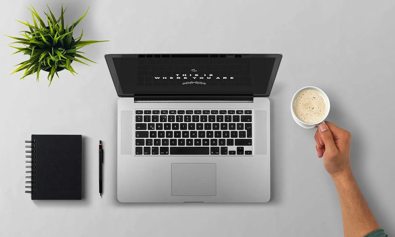 An overhead shot of a laptop, notebook, pen, plant, and a hand holding coffee on a desk.