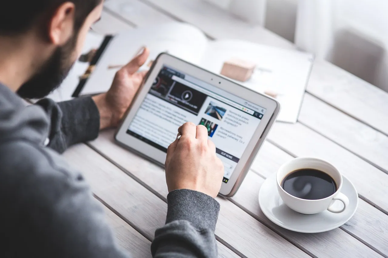 A person in a grey hoodie uses a tablet while sitting at a white wooden table with a cup of coffee.