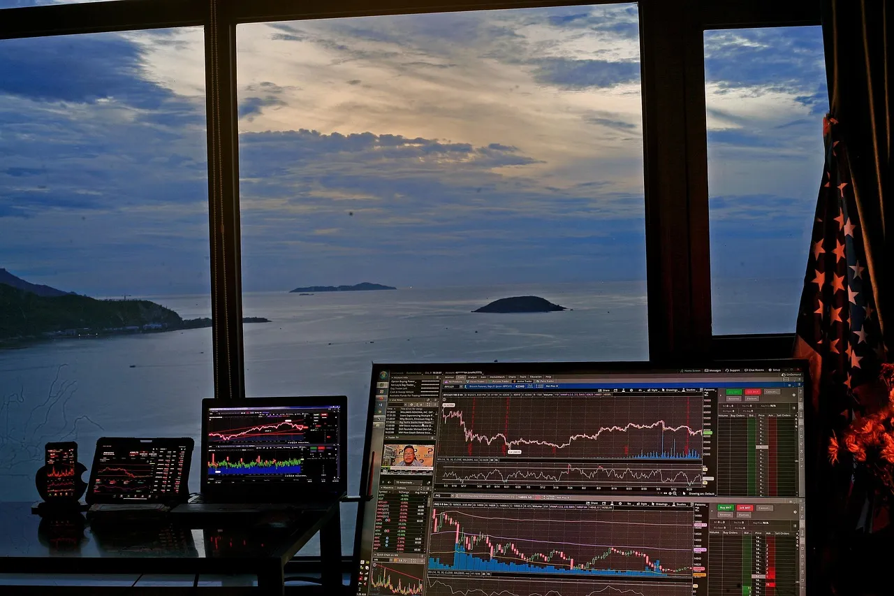 A multi-screen trading setup overlooks a scenic ocean view with islands at dusk.