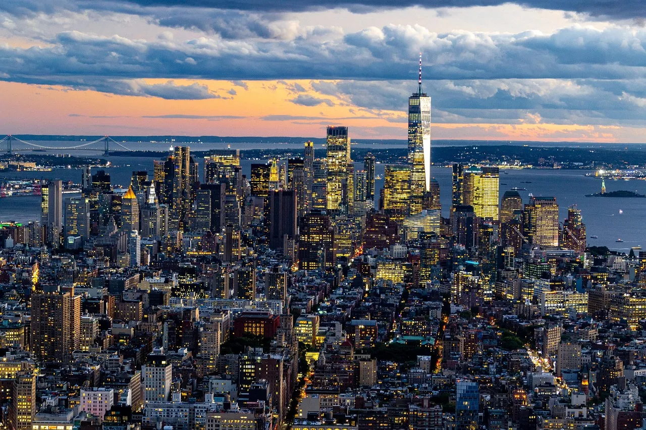 An aerial view of the New York City skyline at dusk, with illuminated buildings and a cloudy sky.
