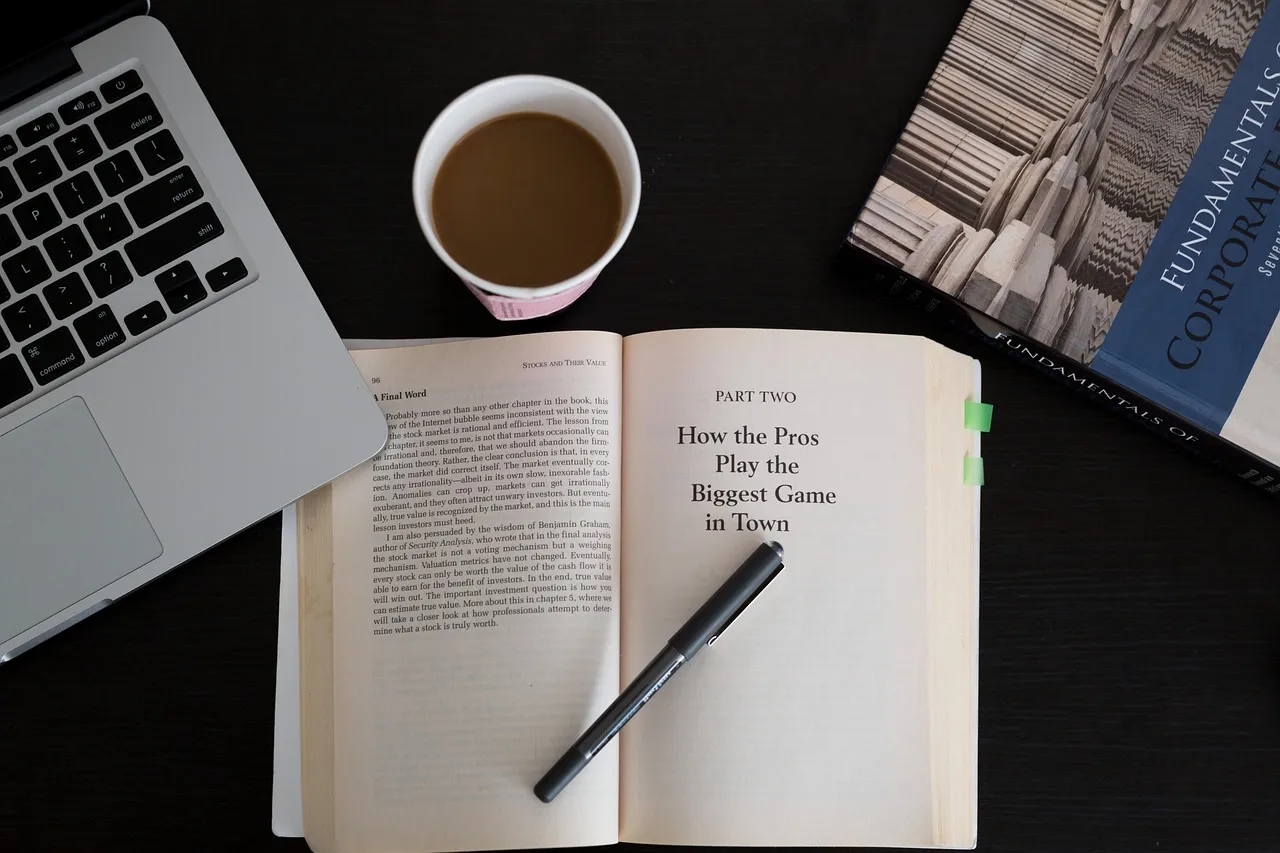 An overhead shot of a laptop, coffee, open book, pen, and another book on a dark surface.