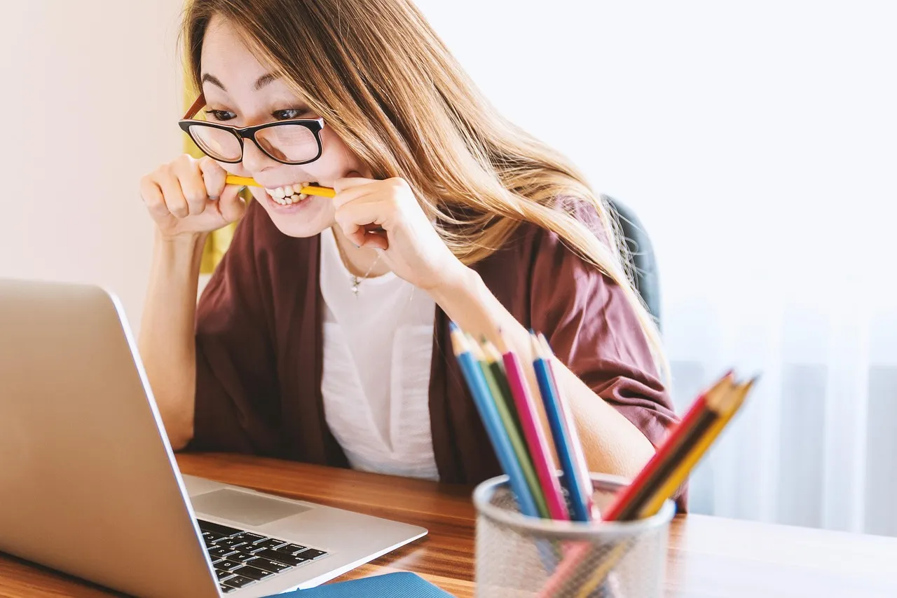 A young woman with glasses bites a pencil while looking intently at a laptop.