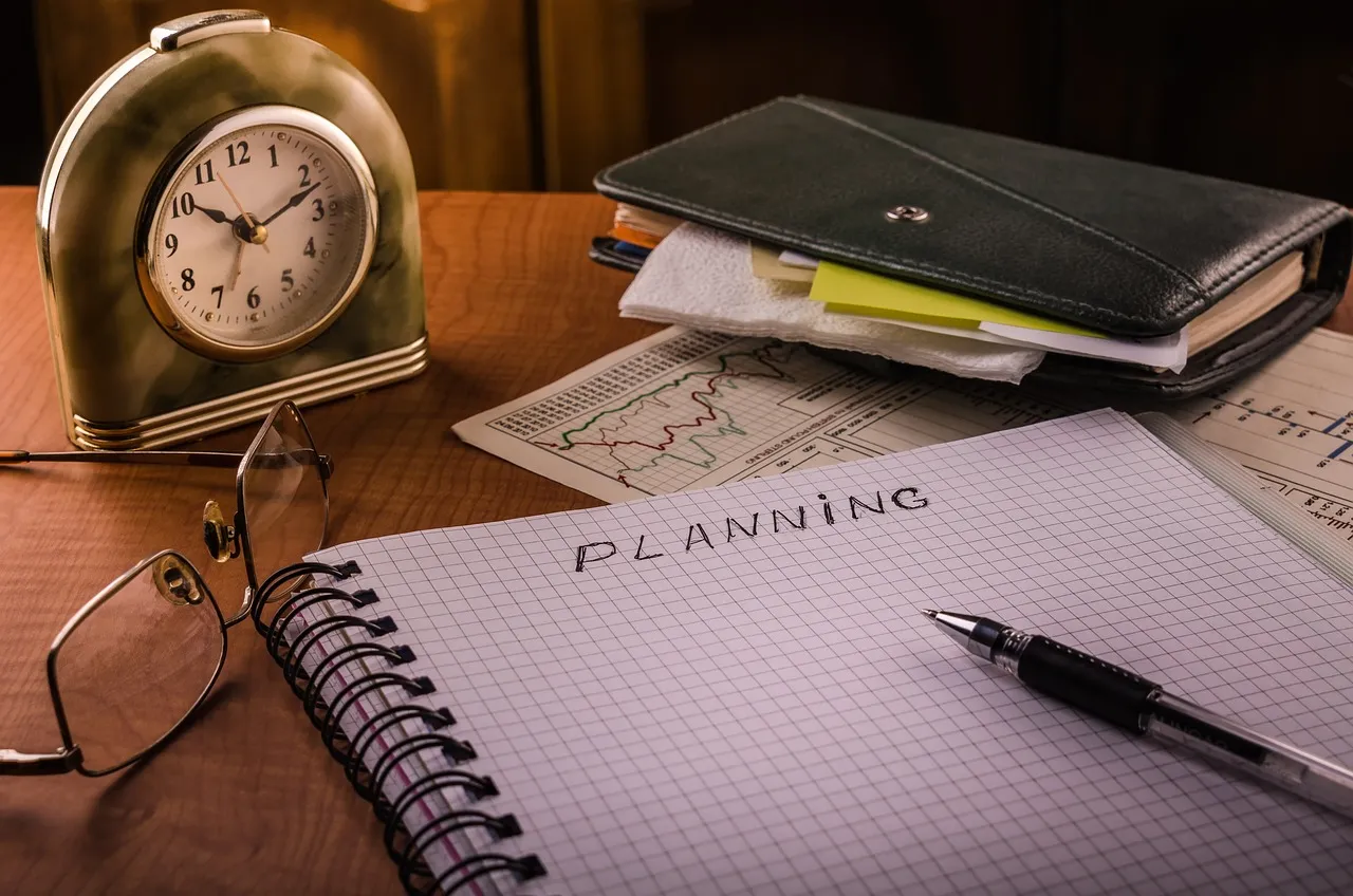 A desk with an alarm clock, glasses, a notebook with "PLANNING" written on it, and financial documents.