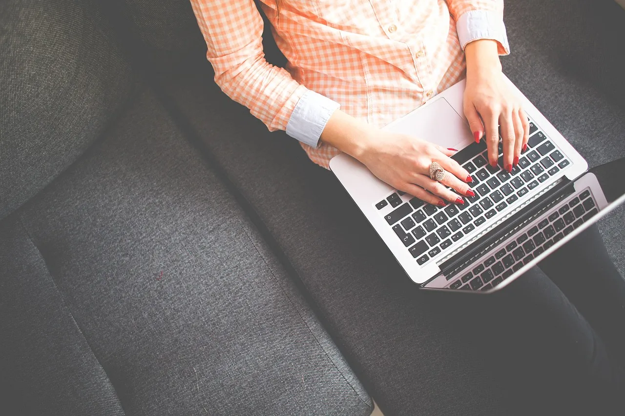 Overhead shot of a person in a plaid shirt typing on a laptop while sitting on a couch.