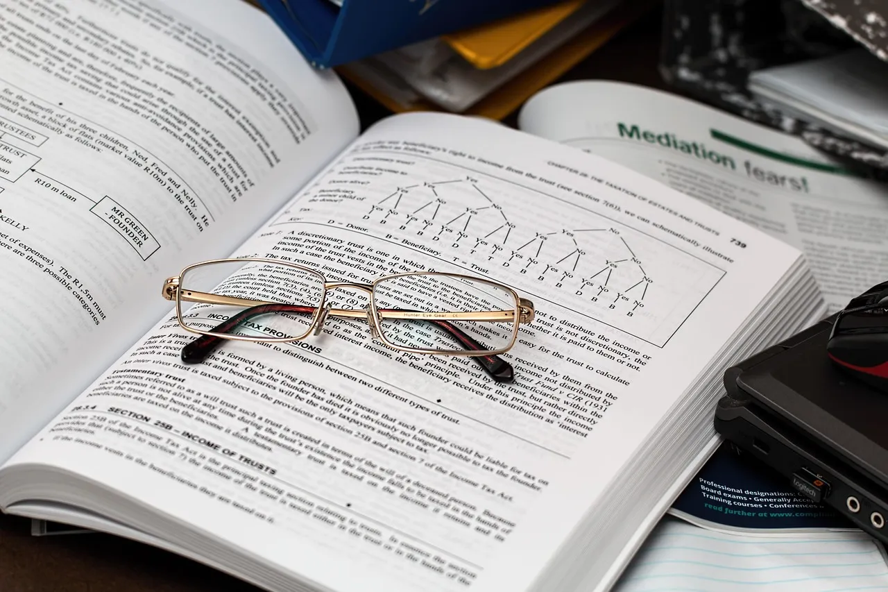 An open textbook with reading glasses resting on it, surrounded by other documents.