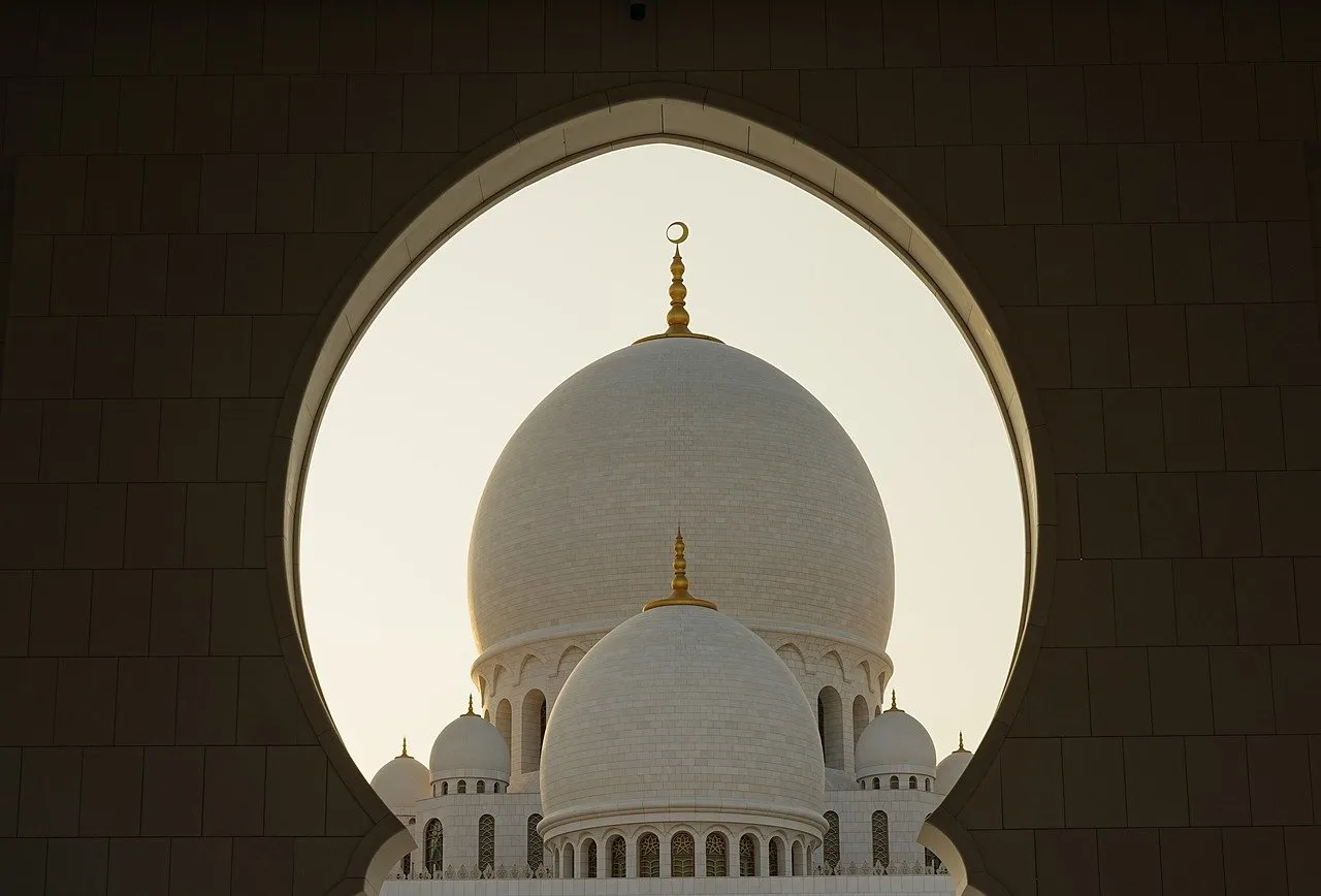 A grand mosque's domes and minarets are framed by an arched entryway against a bright sky.
