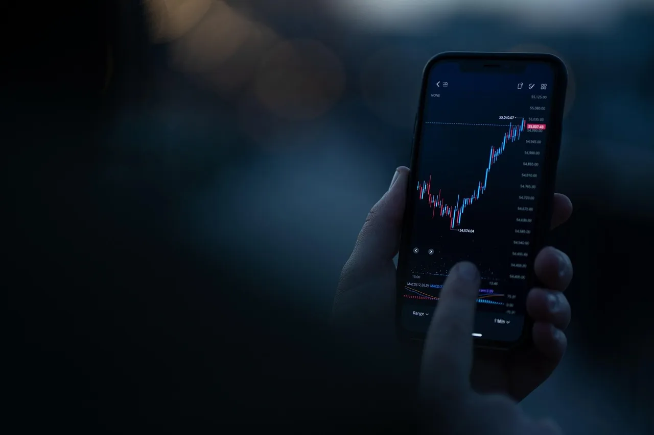 A person's hand holds a smartphone displaying a stock market chart with red and blue lines.