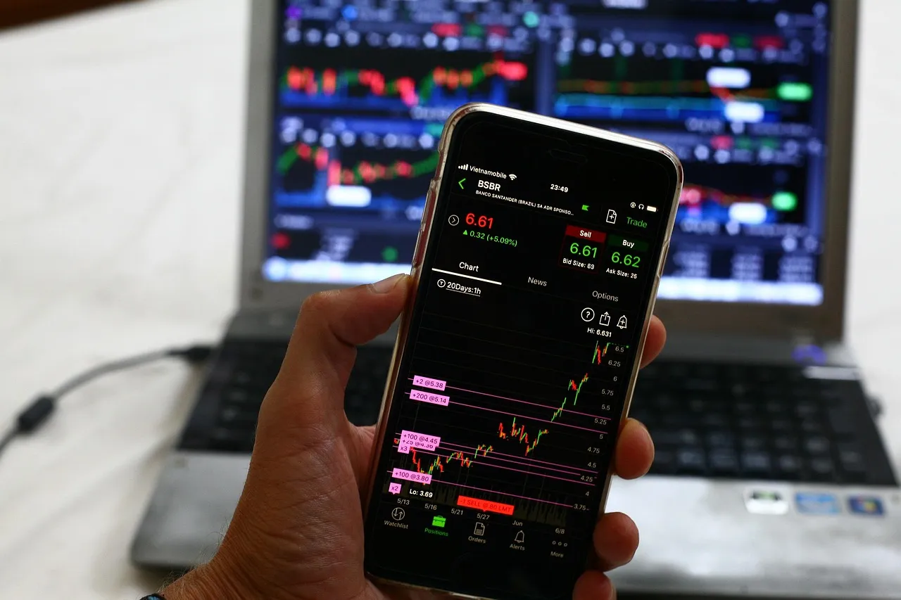 A person holds a smartphone displaying a stock trading app with a laptop showing charts in the background.