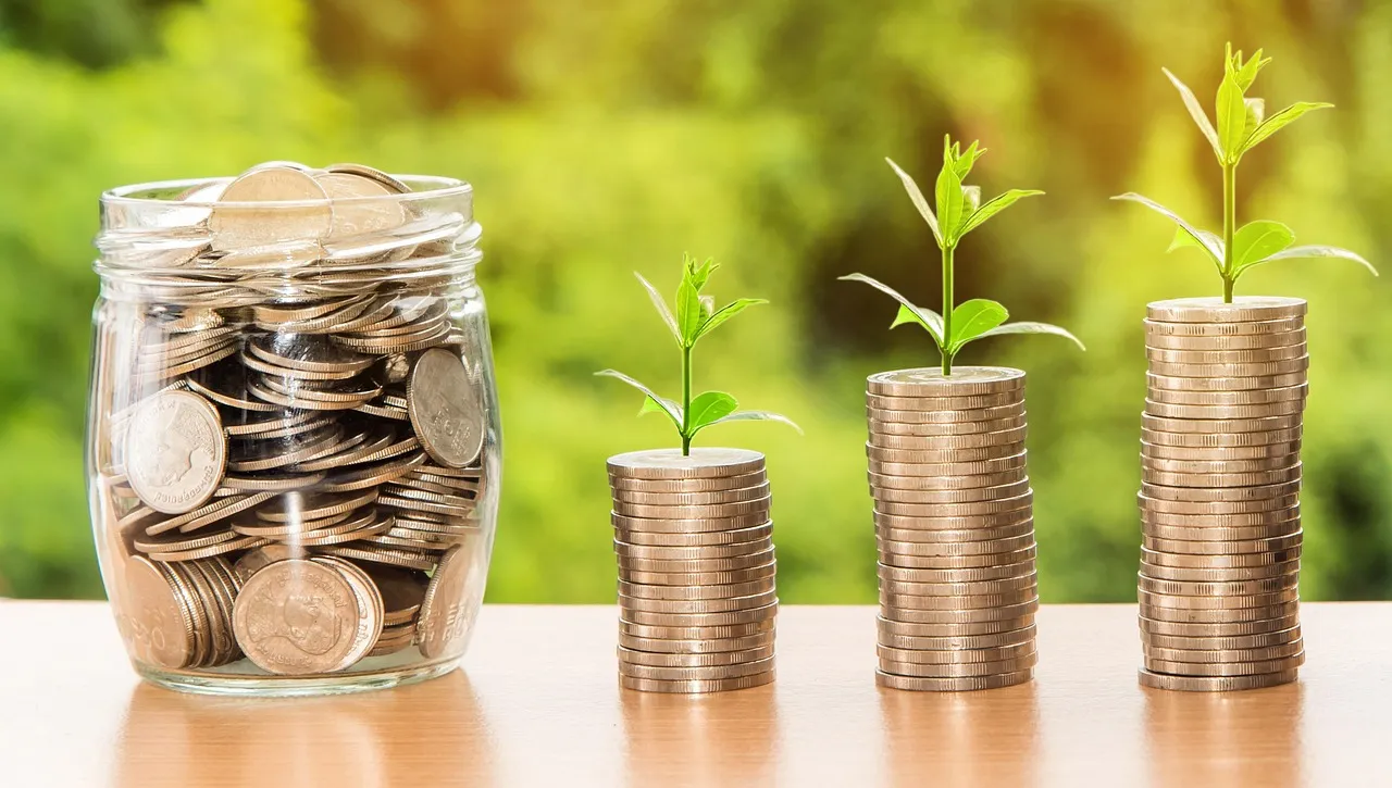 A glass jar full of coins next to three stacks of coins with plants growing from them.