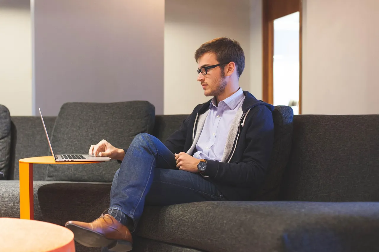 A young man wearing glasses and a hoodie sits on a couch, working on a laptop.