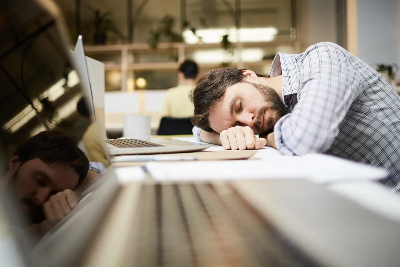 A tired man with a beard is sleeping on his desk in an office setting.