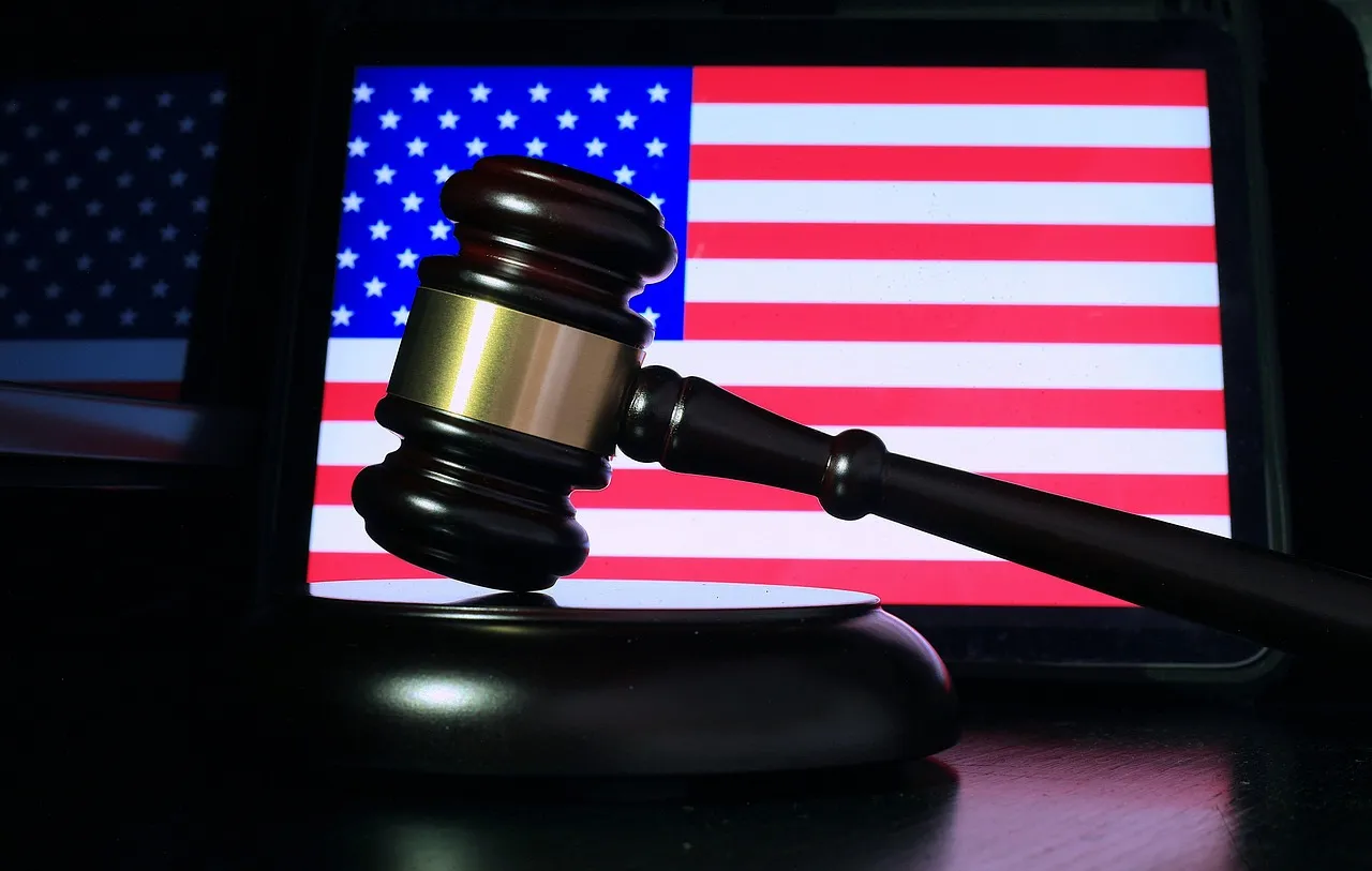 A gavel rests on a dark surface with the American flag displayed on a screen in the background.