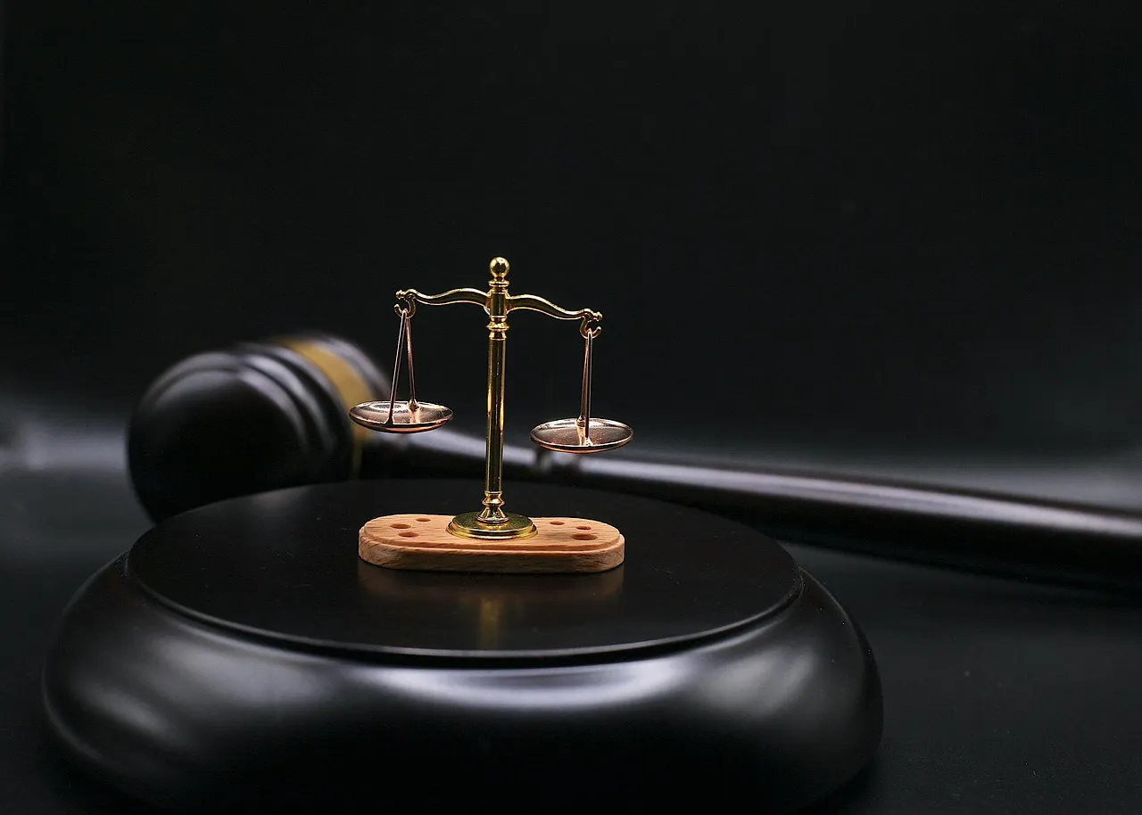 A golden scale of justice sits on a black base with a gavel in the background.
