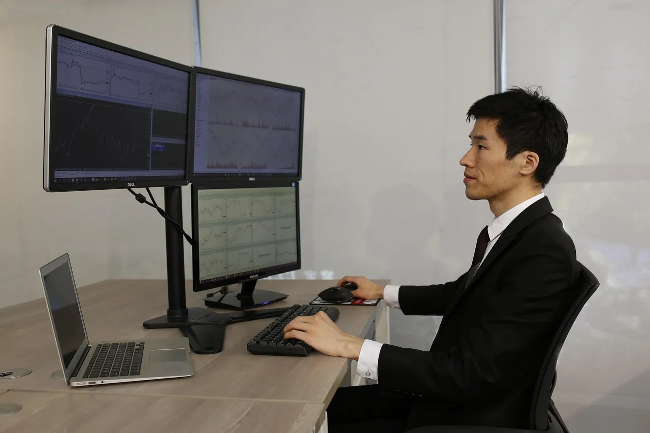 A man in a suit sits at a desk with multiple monitors displaying financial charts.