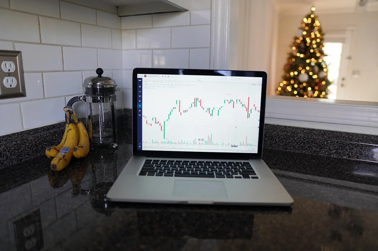 A laptop displaying a stock chart sits on a kitchen counter with bananas and a French press, with a Christmas tree in the background.