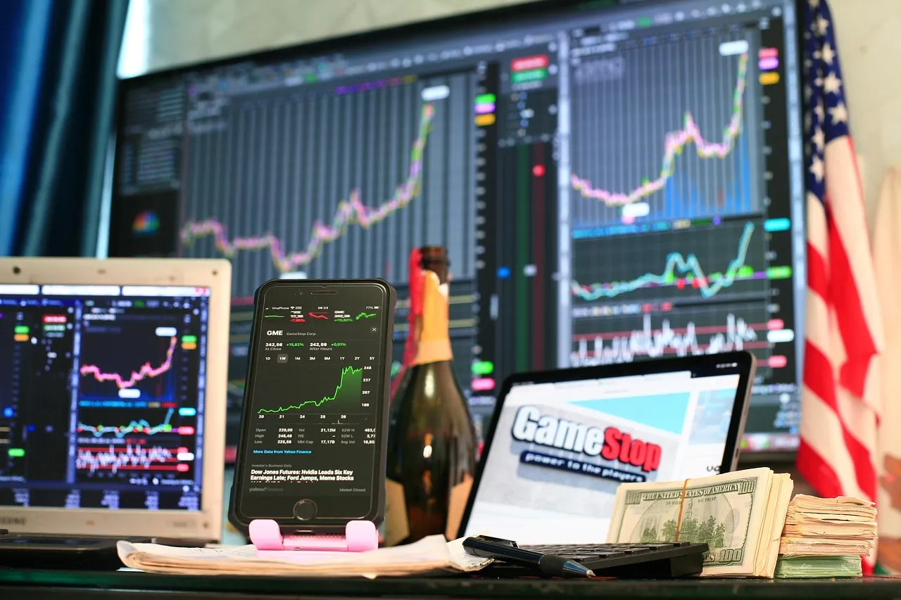 A desk with multiple screens displaying stock charts, a phone showing GME stock, and cash.
