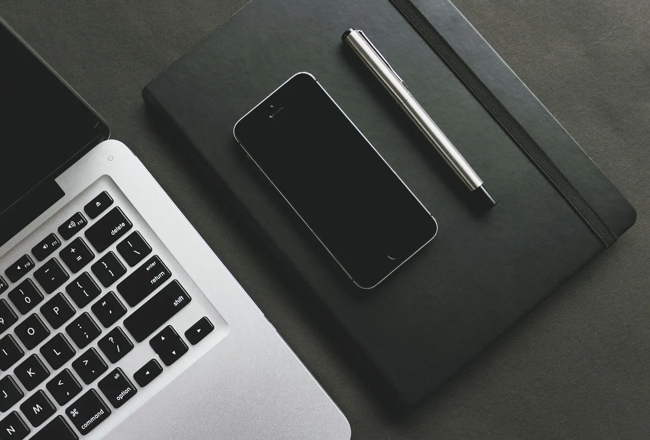A top-down view of a laptop, smartphone, pen, and notebook on a dark surface.