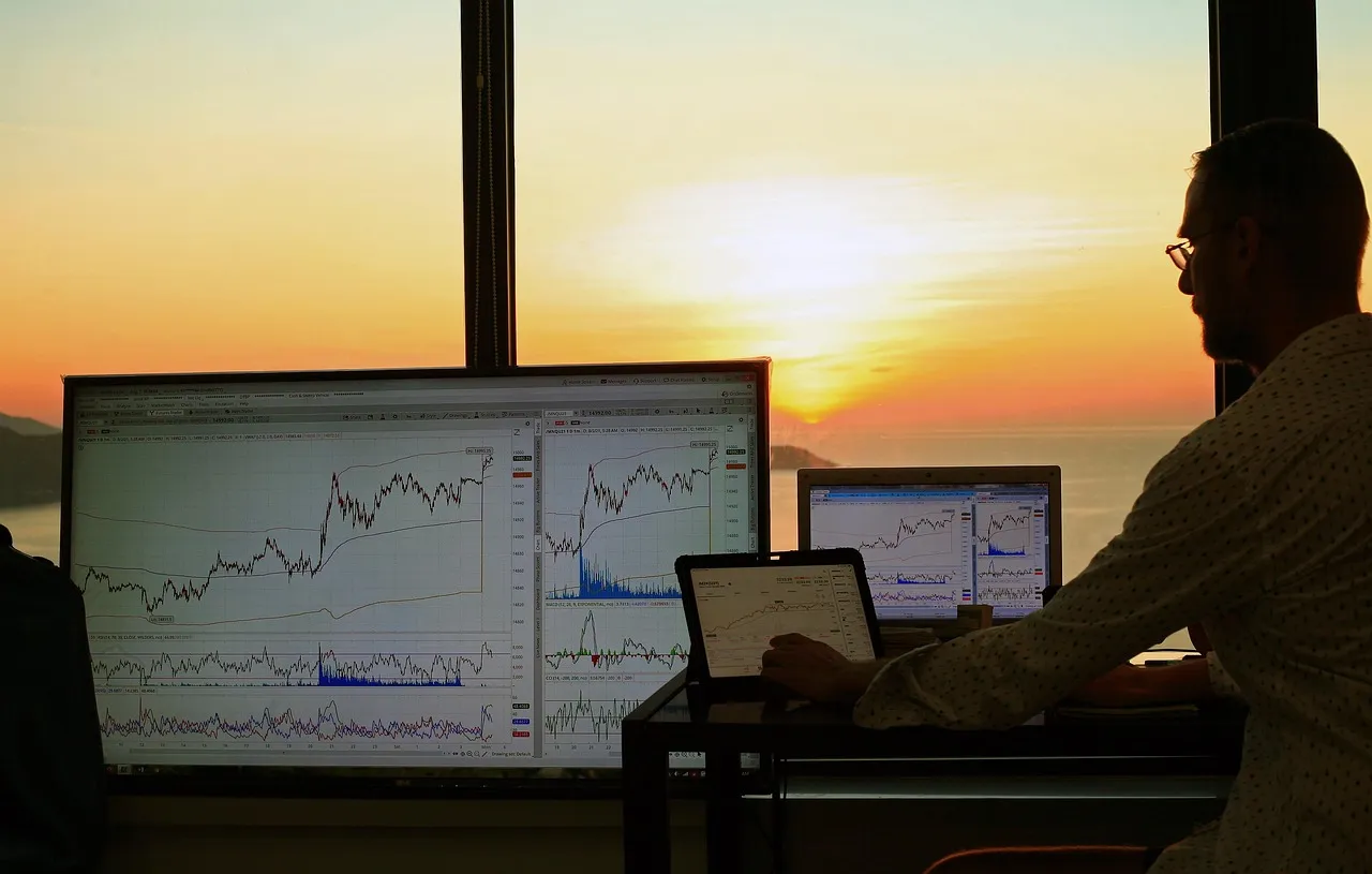 A person monitors multiple screens displaying financial charts against a sunset backdrop.