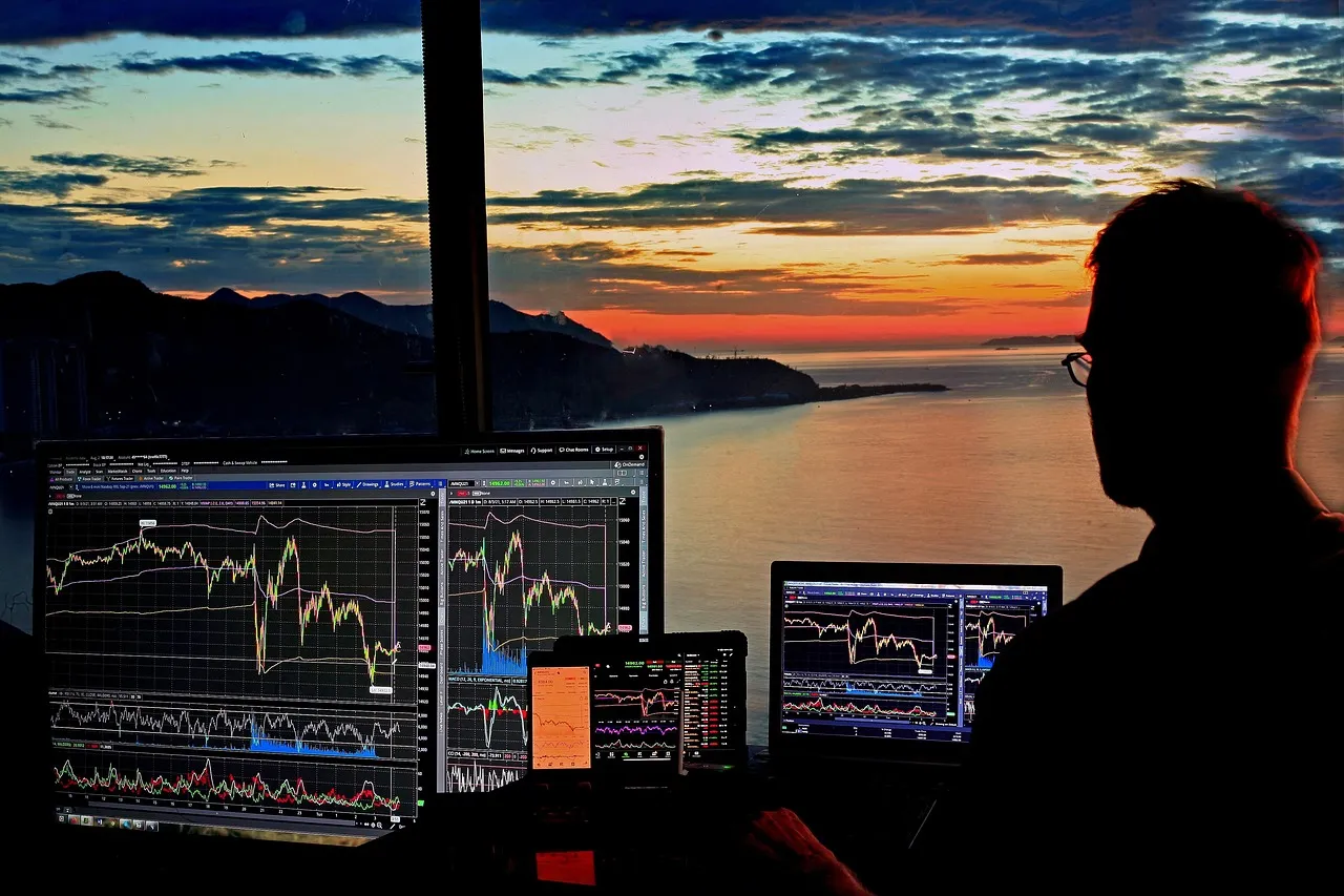 A person monitors multiple screens displaying financial charts against a stunning sunset over the ocean.