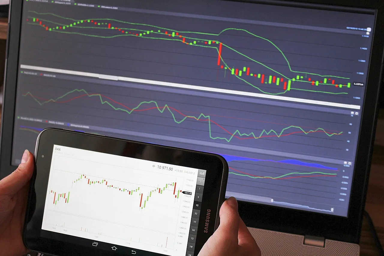 A person holds a tablet displaying stock charts in front of a laptop with more detailed trading graphs.